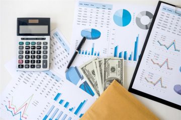 Máy tính cầm tay, cây bút, xấp tiền giấy và phong bì đặt trên các tờ bảng số liệu có biểu đồ cột, biểu đồ tròn
