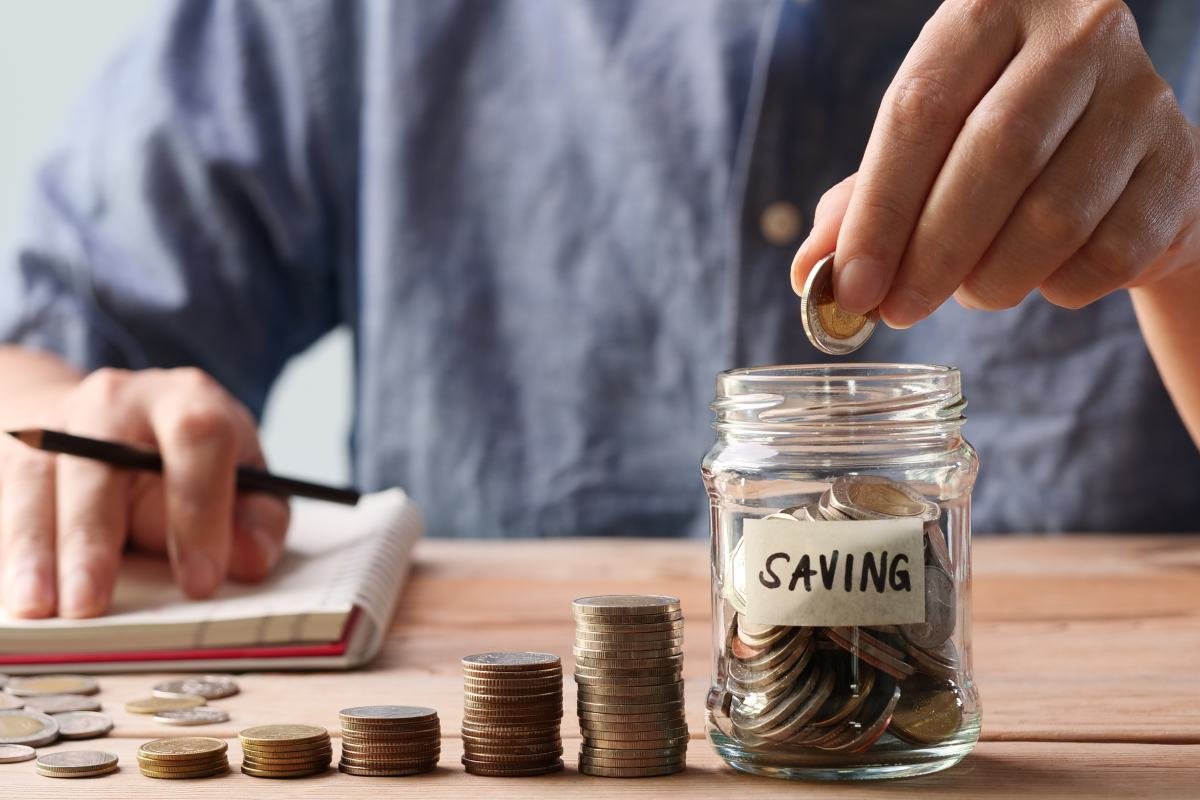 Bàn tay bỏ đồng xu vào hũ thủy tinh dán nhãn SAVING, cạnh các chồng xu và cuốn sổ ghi chép